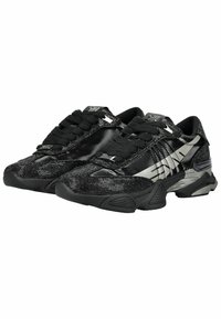 Black and silver low-top sneakers with textured fabric, glossy panels, thick black laces, and sculpted rubber soles.