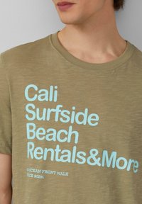 Olivegrünes Baumwoll-T-Shirt mit hellblauem Text, der "Cali Surfside Beach Rentals & More" lautet, mit runder Halslinie und kurzen Ärmeln.