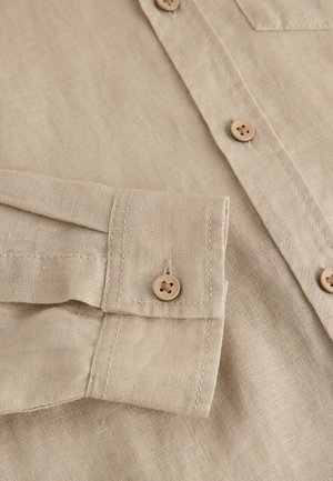 Beige Leinenhemd im Nahaufnahme mit leichter Textur, braunen Knöpfen und einer einzelnen Brusttasche. Verfügt über eine Manschette mit Knopfdetail und Naht.