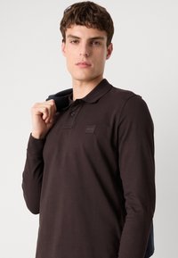Mørkebrun langærmet polo-shirt med klassisk krave, der har et logo-patch på brystet og et blødt, struktureret stof.