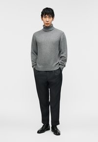 Νεαρός άνδρας όρθιος, φορώντας γκρι πουλόβερ με ψηλό γιακά, σκούρο παντελόνι με λεπτές ρίγες και μανσέτες, και μαύρα δερμάτινα επίσημα παπούτσια, με τα χέρια στις τσέπες.