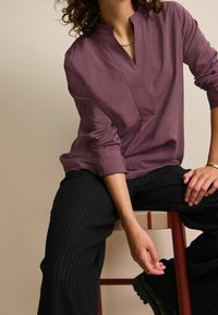 Top a maniche lunghe color prugna con orlo asimmetrico e scollo a V, abbinato a pantaloni neri a righe sottili e scarpe basse nere.