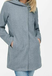 Abrigo gris con capucha fabricado con tejido texturizado, de corte recto, con bolsillos laterales y diseño minimalista sin cierres visibles.