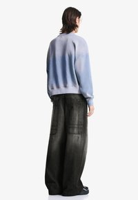 O persoană cu părul până la umeri poartă un pulover albastru decolorat și blugi negri largi cu buzunare mari la spate, văzută din spate.