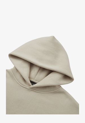 Bež dukserica s velikom, mekanom kapuljačom, rebrastim okovratnikom i bešavnim dizajnom. Tkanina izgleda glatko i blago rastezljivo. Oznaka s logotipom vidljiva je iznutra.