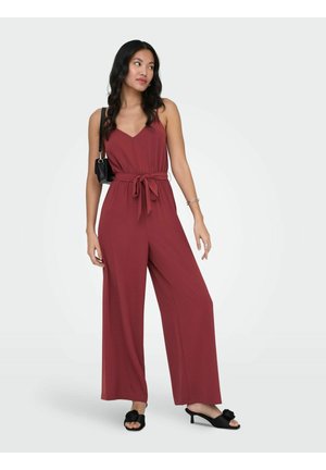 Kvinne iført en ermeløs, rustrød vidbukse-jumpsuit med knyttet midje, svarte sandaler med hæl, og en svart skulderveske.