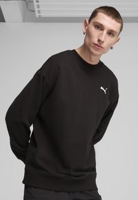 Sudadera negra hecha de una tela suave, con cuello redondo, puños acanalados y un logo blanco en el pecho. Diseño sencillo y minimalista.