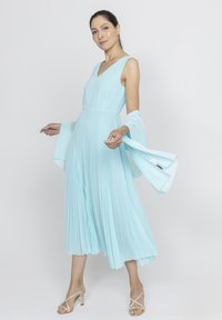 Jasnoniebieska, plisowana sukienka o dekolcie w kształcie litery V i zwiewnych rękawach, z dopasowaną górą i długością do połowy łydki. Noszona z sandałkami na obcasie.