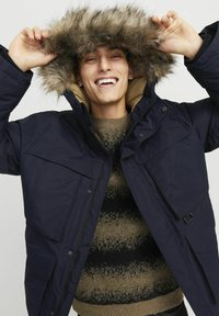 Marineblauwe parka jas met een bontgevoerde capuchon, voorzien van voorzakken, een ritssluiting en een beige binnenvoering, gedragen over een gestreepte trui.