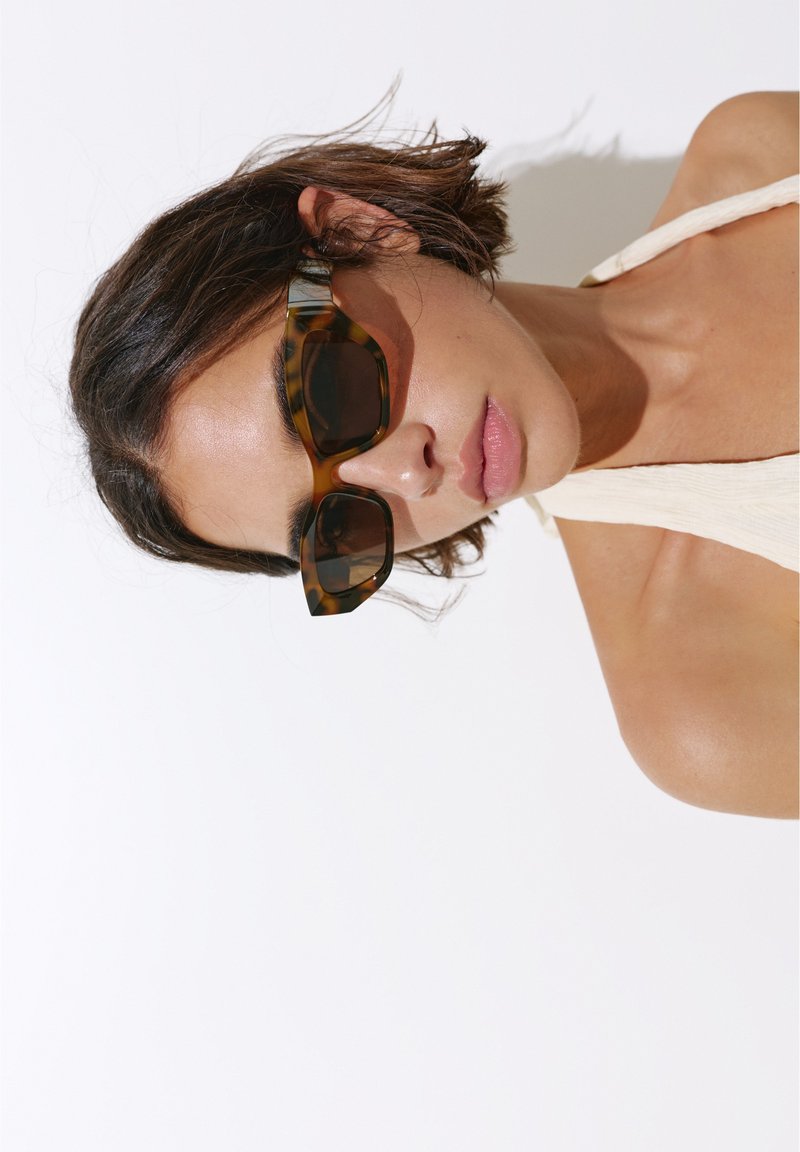 Femme aux cheveux courts et foncés portant des lunettes de soleil écaille de tortue marron et un haut clair sans manches sur fond blanc.