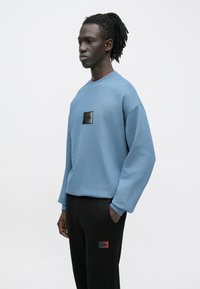 Fiatal férfi sötét dreadlock hajjal, kék pulóvert és fekete nadrágot viselve, egy kézzel a zsebében áll egyszínű háttér előtt.