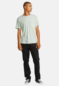 Lichtgroene kraagloze t-shirt met korte mouwen en een borstzak, gecombineerd met zwarte broek en zwarte sneakers met witte zolen.