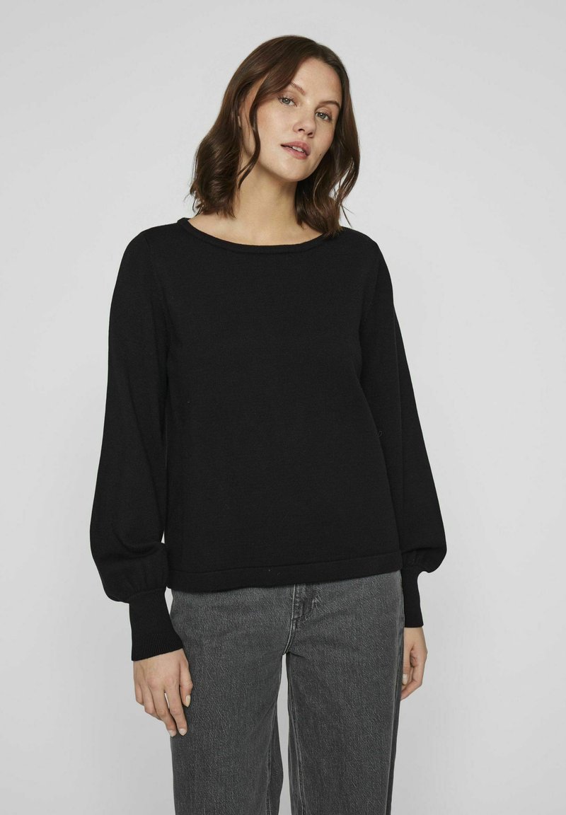 Pull noir à encolure ronde, manches longues bouffantes, poignets côtelés et coupe décontractée, associé à un pantalon gris taille haute.