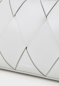 Harmaa nahkalaukku, jossa on punottu muotoilu, sileä pinta ja huomaamaton Calvin Klein -logo oikeassa alakulmassa.