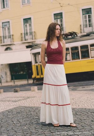 Young woman in burgundy sleeveless top and white skirt with red trim stands on cobblestone street near yellow tram.