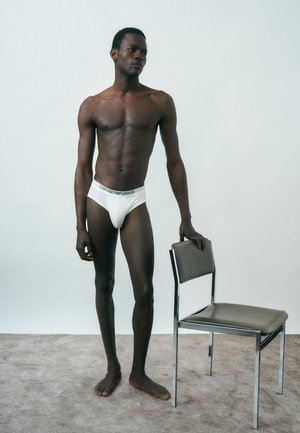 Slank mørk mand iført hvide Emporio Armani underbukser, stående barfodet på tæppe ved siden af en metalstol med grå pude, mod ensfarvet baggrund.