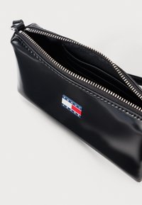 Pochette en cuir noir avec une fermeture éclair argentée, ouverture sur le dessus montrant les compartiments intérieurs, et un petit logo rectangulaire Tommy Jeans.