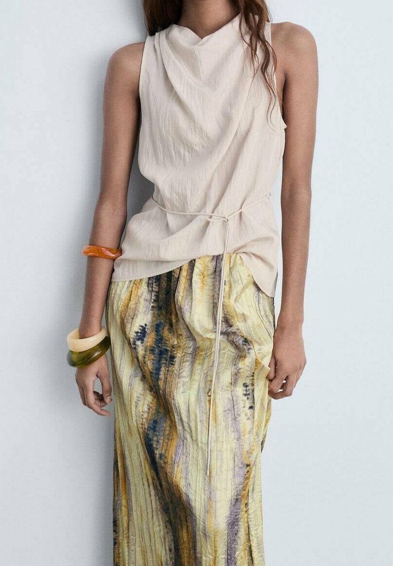 Top beige senza maniche con scollatura drappeggiata, abbinato a una lunga gonna a fantasia nei toni del giallo, blu e marrone. Accentuato da vari bracciali.