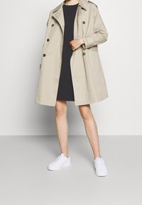 Beige trenchcoat med dubbelknäppt design, stora knappar och axelklaffar, kombinerad med en svart klänning och vita sneakers. Slät textur.