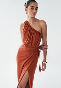 Jednodelné šaty v rezavě oranžové barvě vyrobené z jemné látky; mají zavinovací korzet, vysoký rozparek a vázací detail v pase.