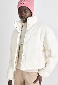 Veste matelassée en velours côtelé blanc avec boutons pression et patch logo, superposée à un col roulé beige. Bonnet en tricot rose sur la tête du modèle.