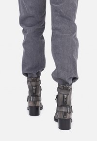 Botas de cuero negro hasta el tobillo con un tacón grueso, que cuentan con múltiples cremalleras y correas, combinadas con joggers texturizados grises.