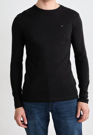 Schwarzes Langarmshirt aus weichem Material mit rundem Ausschnitt und dezentem Logo auf der Brust. Kombiniert mit blauen Jeans.
