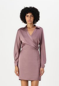 Robe enveloppante en satin mauve, avec des manches longues et un col en V. Elle présente une taille cintrée et un drapé subtil, se terminant au-dessus du genou.