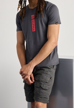 T-shirt gris à manches courtes avec un graphique rouge "ALPHA" sur le devant, associé à un short cargo de couleur gris foncé avec plusieurs poches.