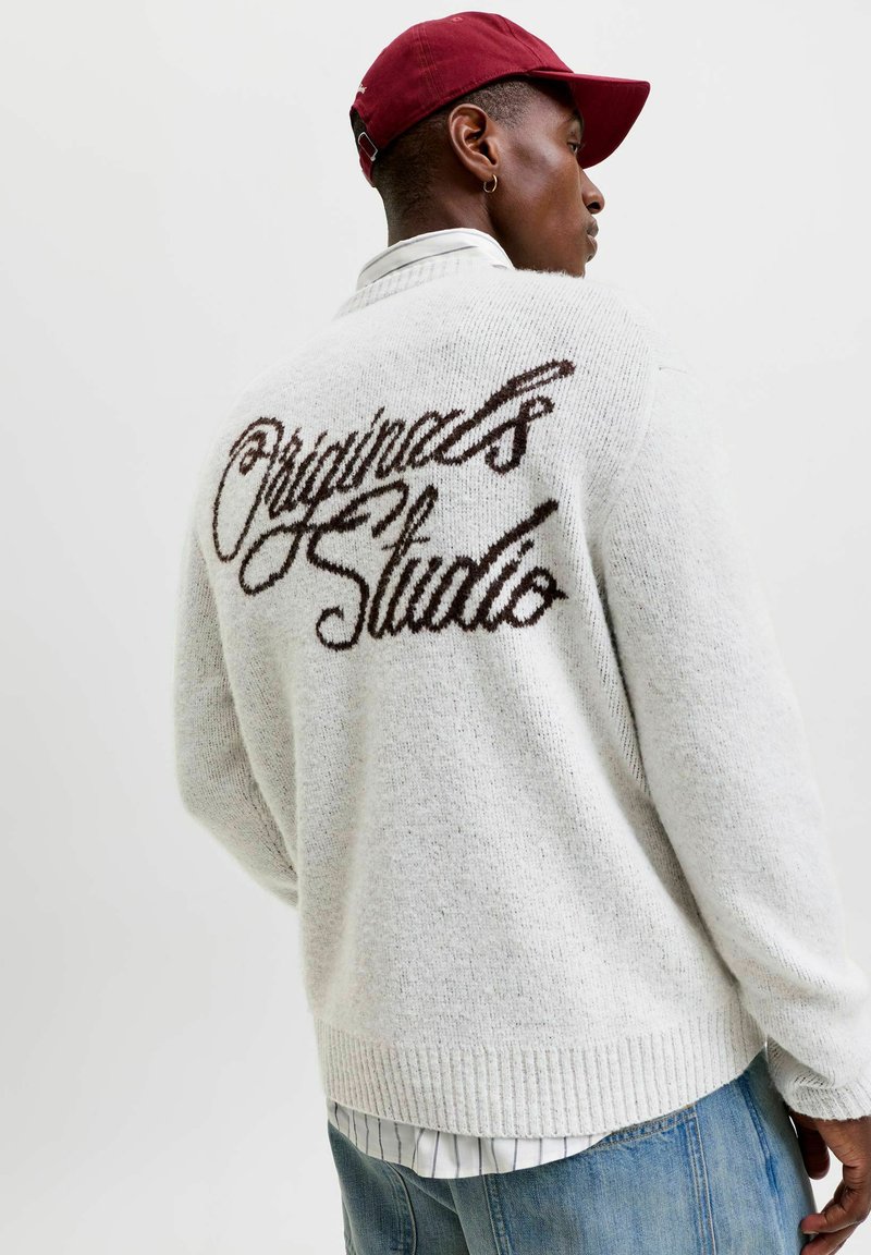 Personne portant une casquette rouge, une chemise blanche et un pull clair avec "Originals Studio" en écriture manuscrite dans le dos, tournée dos à la caméra.