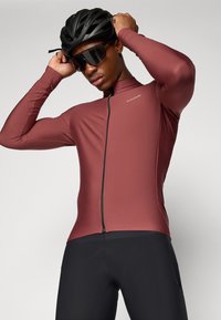 Bordeaux kleurig fietsshirt met lange mouwen en een rits aan de voorkant, gemaakt van rekbaar materiaal. Draagt met zwarte fietsshorts en een helm.