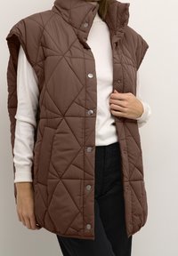 Brązowa pikowana kamizelka z wysokim kołnierzem, z zatrzaskami i bocznymi kieszeniami, noszona na białej bluzce z długim rękawem.