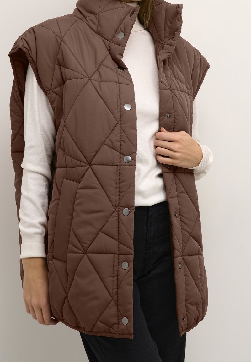 Brązowa pikowana kamizelka z wysokim kołnierzem, z zatrzaskami i bocznymi kieszeniami, noszona na białej bluzce z długim rękawem.