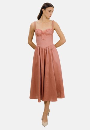 Kvinne iført ermeløs midi-kjole i støvet rosa med flettede stropper og sløyfdetalj på tilpasset overdel, kombinert med beige hæler, stående på hvit bakgrunn.