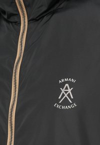 Juodas džemperis su smėlio spalvos užtrauktuku ir baltu Armani Exchange logotipu ant krūtinės.