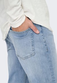 Jeans de mezclilla azul claro con un ajuste relajado, que cuentan con un bolsillo trasero y un acabado limpio. Llevados con una camisa de manga larga color crema.