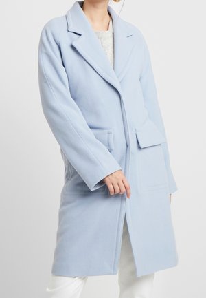 Femme portant un manteau bleu clair longueur genou avec de larges revers, poches à rabat et manches longues sur un pantalon blanc et un pull clair.