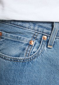 Un jean en denim bleu clair présente un design classique à cinq poches avec des rivets couleur cuivre, des coutures contrastantes et une texture lisse.