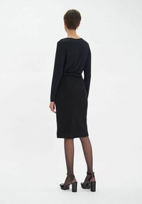 Haut noir à manches longues associé à une jupe noire moulante. La tenue présente une ceinture texturée et des collants noirs transparents avec des sandales à talons.