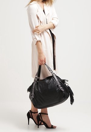 Nainen vaaleassa trenssitakissa ja mustassa mekossa, joka kantaa suurta mustaa nahkalaukkua, jossa on hapsukoristelu, ja käyttää mustia korkeakorkoisia sandaaleja.