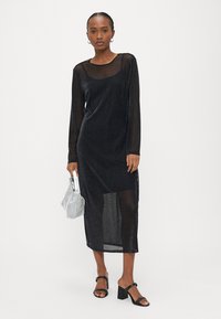 Γυναίκα που φοράει μαύρο διαφανές midi φόρεμα με μακριά μανίκια πάνω από μαύρο φανελάκι, κρατώντας ασημένια τσάντα και φορώντας μαύρα σανδάλια με τακούνια.