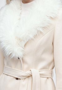 Femme portant un manteau crème ceinturé avec un grand col en fourrure blanche et duveteuse par-dessus un pull blanc.