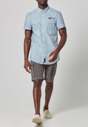 Mężczyzna idący w jasnoniebieskiej koszuli z krótkim rękawem na guziki oraz szarych szortach w kratę z beżowymi butami wsuwanymi.