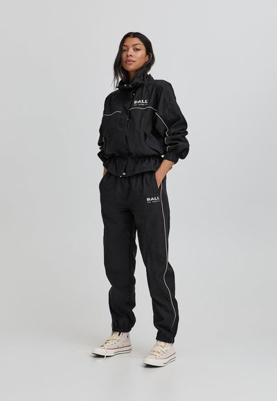 Schwarze Windjacke mit elastischen Bündchen, weißer Paspelierung und Frontreißverschluss. Kombiniert mit passenden Jogginghosen mit elastischem Bund und Logo.