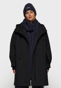 Zwarte parka met capuchon, voorzien van een rits en drukknoopsluiting. Vergezeld van een donkerblauwe wollen sjaal en een bijpassende beanie.