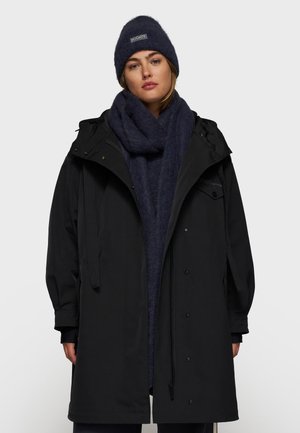 Parka nera con cappuccio, dotata di chiusura con zip e bottoni a pressione. Abbinata a una sciarpa di lana blu scuro e un berretto coordinato.