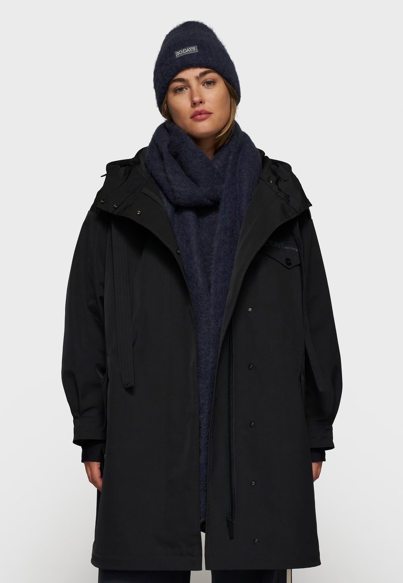 Zwarte parka met capuchon, voorzien van een rits en drukknoopsluiting. Vergezeld van een donkerblauwe wollen sjaal en een bijpassende beanie.
