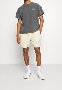 Harmaa puuvillapaita, jossa on lyhyet hihat ja rintalogo, yhdistettynä vaaleanbeigeihin pellavashortseihin. Valkoiset lenkkarit harmailla yksityiskohdilla viimeistelevät asun.