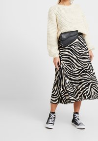 Flauschiger, cremefarbiger Strickpullover, schwarz-weiße, zebra gestreifte Plissee-Rock, schwarze Umhängetasche und schwarze High-Top-Sneaker.
