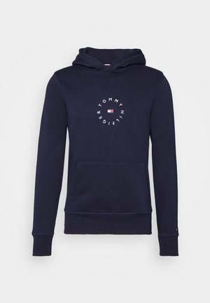 Sweat à capuche bleu marine avec poche avant et texte blanc circulaire "Tommy Hilfiger" entourant le logo du drapeau de la marque en rouge, blanc et bleu.